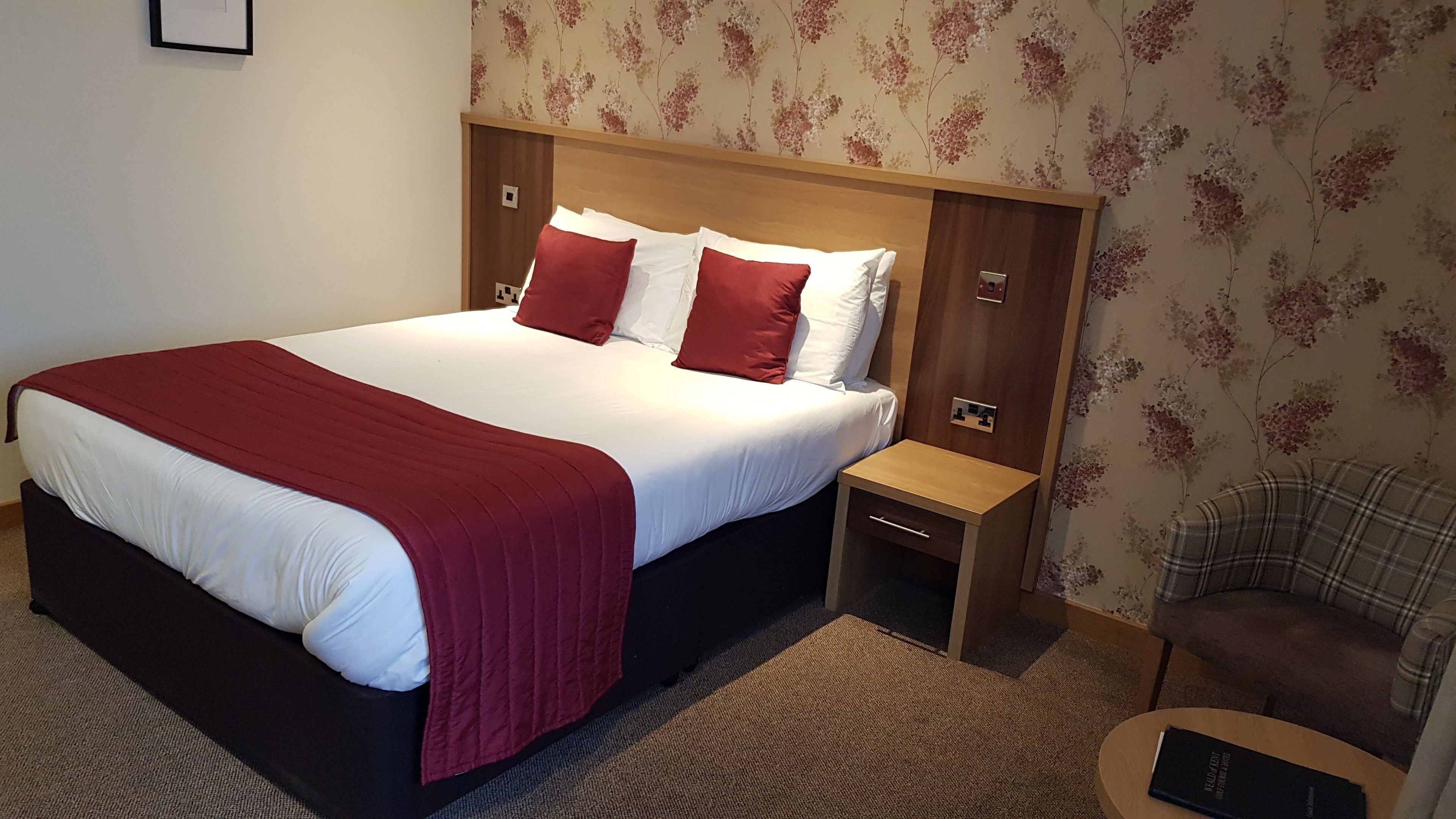 A neatly made bed with a red blanket, a wooden headboard, and a nightstand with a lamp.