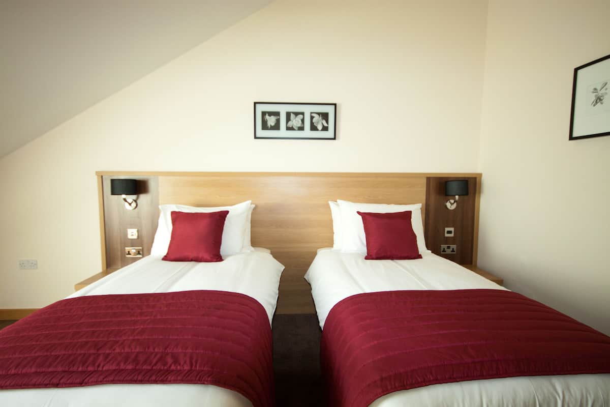 Habitación de hotel con dos camas, cada una con colcha roja y ropa de cama blanca. Hay lámparas de pared a ambos lados de las camas, y un cuadro enmarcado encima.