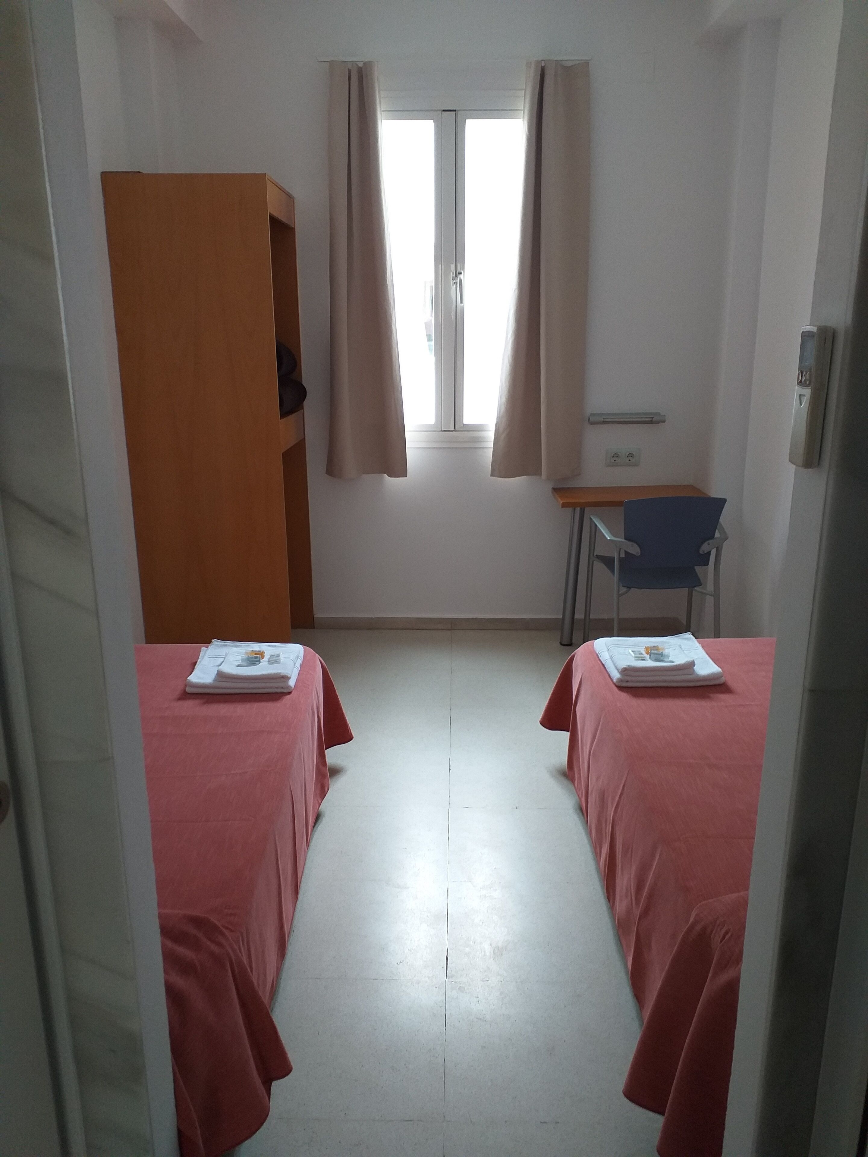 A room with two beds, a desk, a chair, a wardrobe, and a window with curtains.