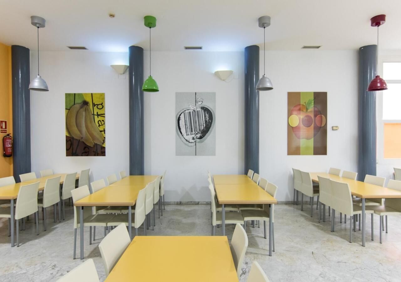 Un comedor con mesas y sillas amarillas, lámparas colgantes verdes y rojas, y obras de arte en las paredes.