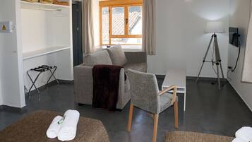 Habitación de hotel con sofá, dos sillas, una mesita, una lámpara de pie, una ventana con cortinas y un perchero para toallas.