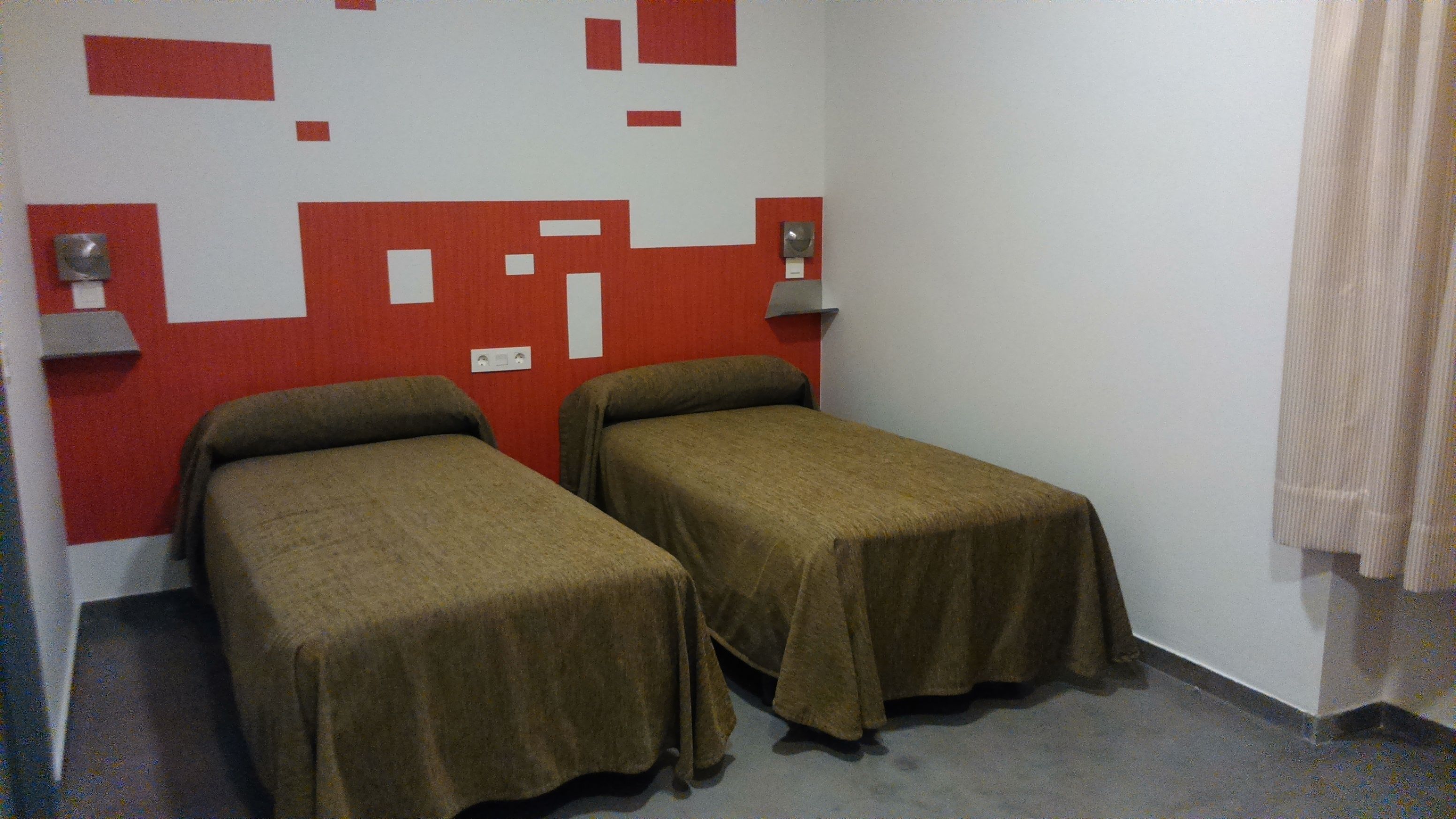 Une chambre avec deux lits simples, un décor mural aux couleurs rouge et blanche, et un sol gris.