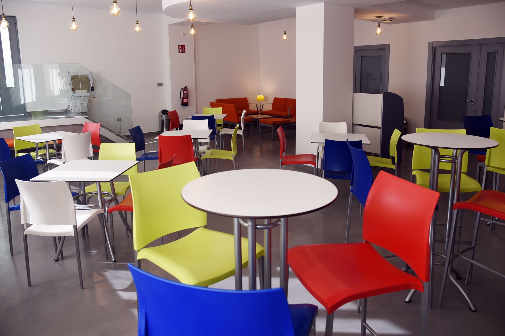 Un espace de restauration moderne avec des chaises et des tables aux couleurs vives, un extincteur accroché au mur et un escalier en arrière-plan.