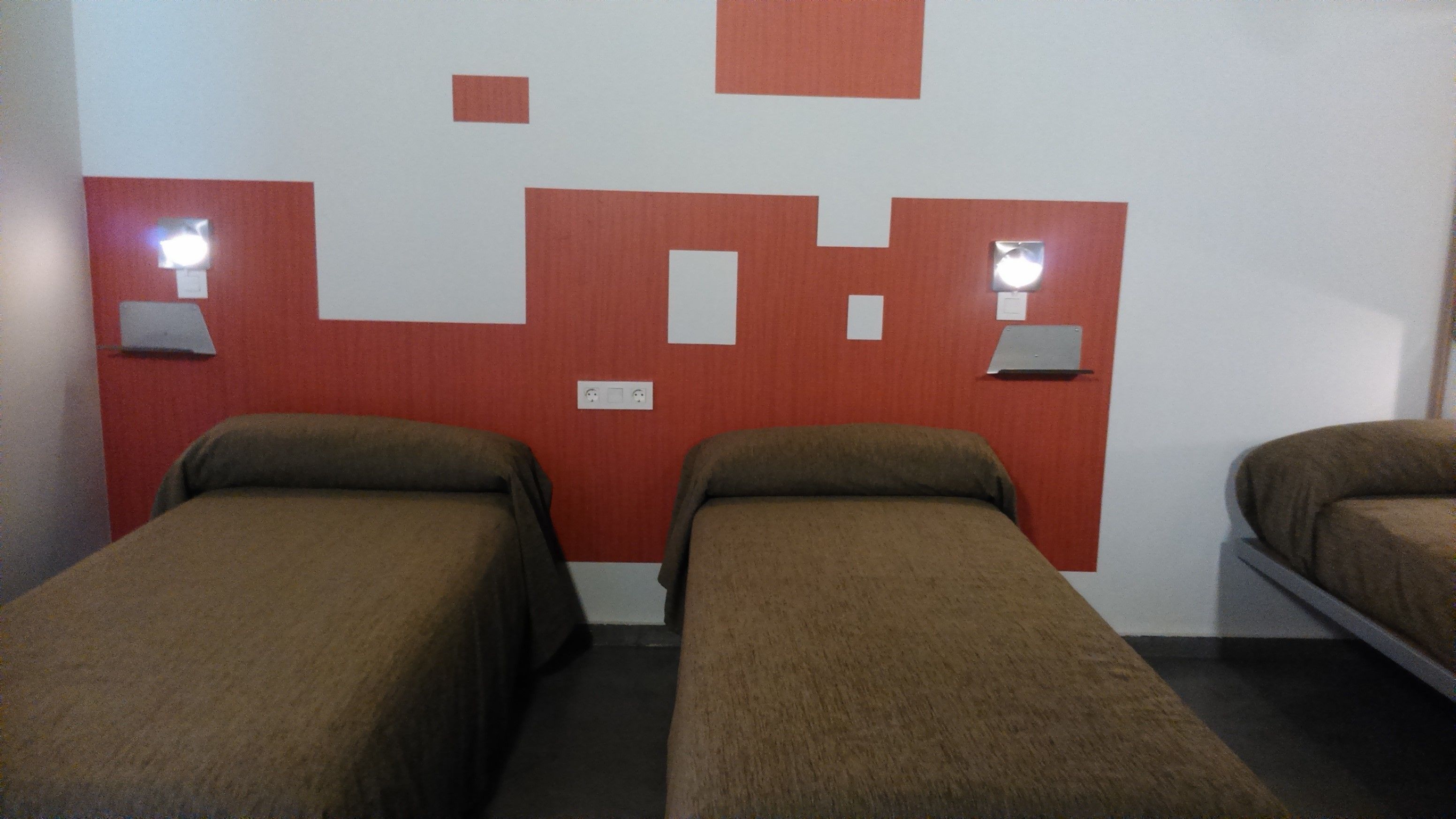 A room with two single beds, each with a brown bedspread. There are two wall-mounted lights on either side of the beds. The wall has a red and white geometric pattern.