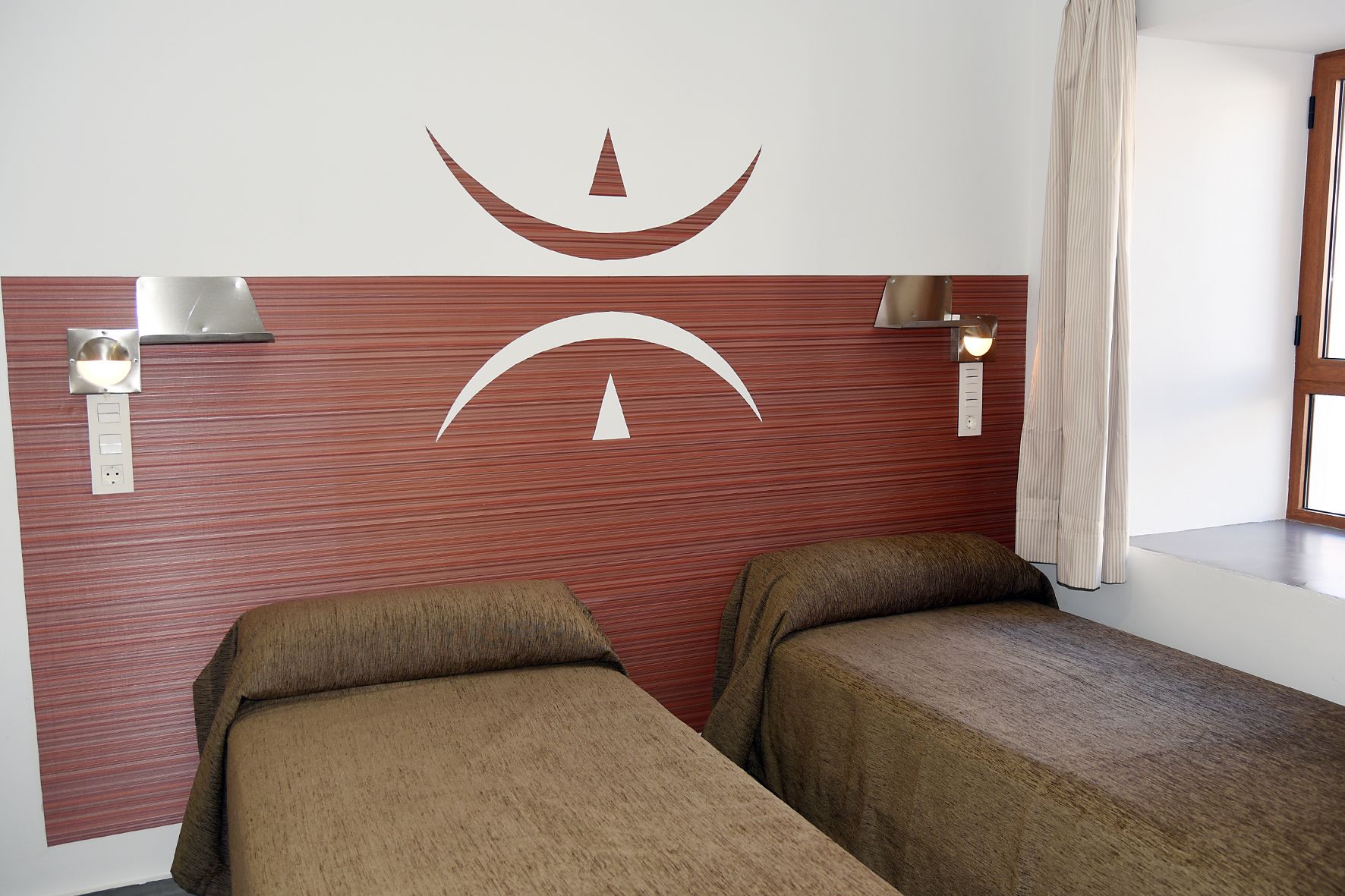Habitación de hotel con dos camas individuales, cabecero de madera, lámparas de pared y una ventana con cortinas.