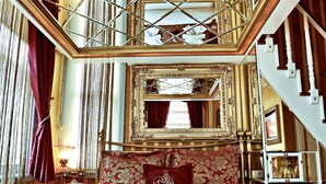Ein luxuriöses Schlafzimmer mit einem großen Bett, einem Kopfteil in Rot und Gold, einem großen Spiegel und einer Treppe zu einer oberen Ebene.