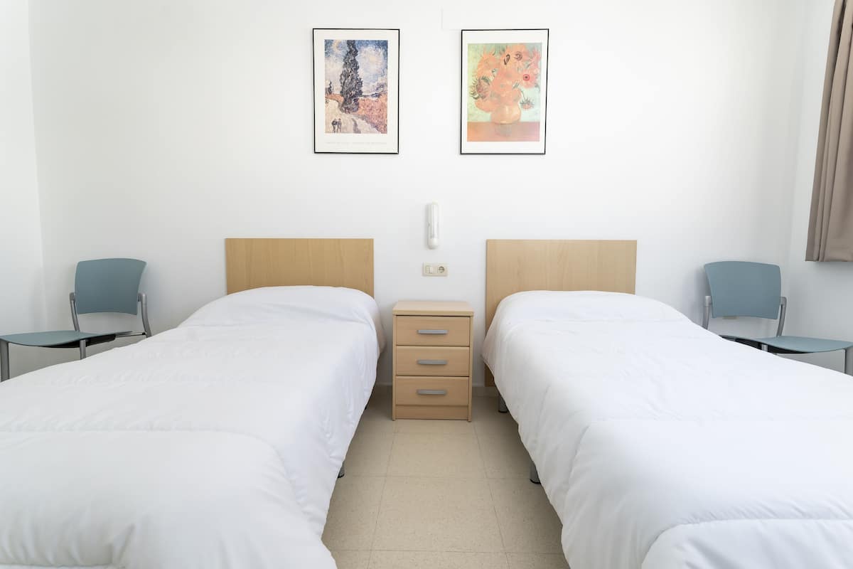 A hotel room with two single beds, a small bedside table, a chair, and two framed artworks on the wall.