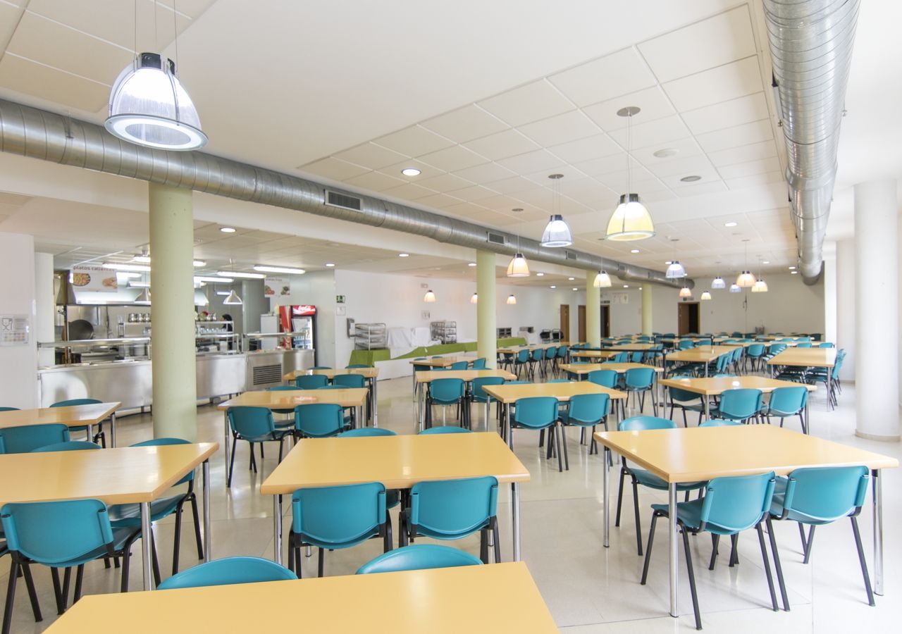 A cafeteria with multiple tables and chairs, a kitchen area in the background, and ventilation ducts on the ceiling.