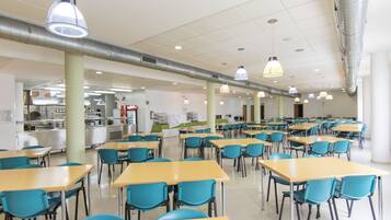 A cafeteria with multiple tables and chairs, a kitchen area in the background, and ventilation ducts on the ceiling.
