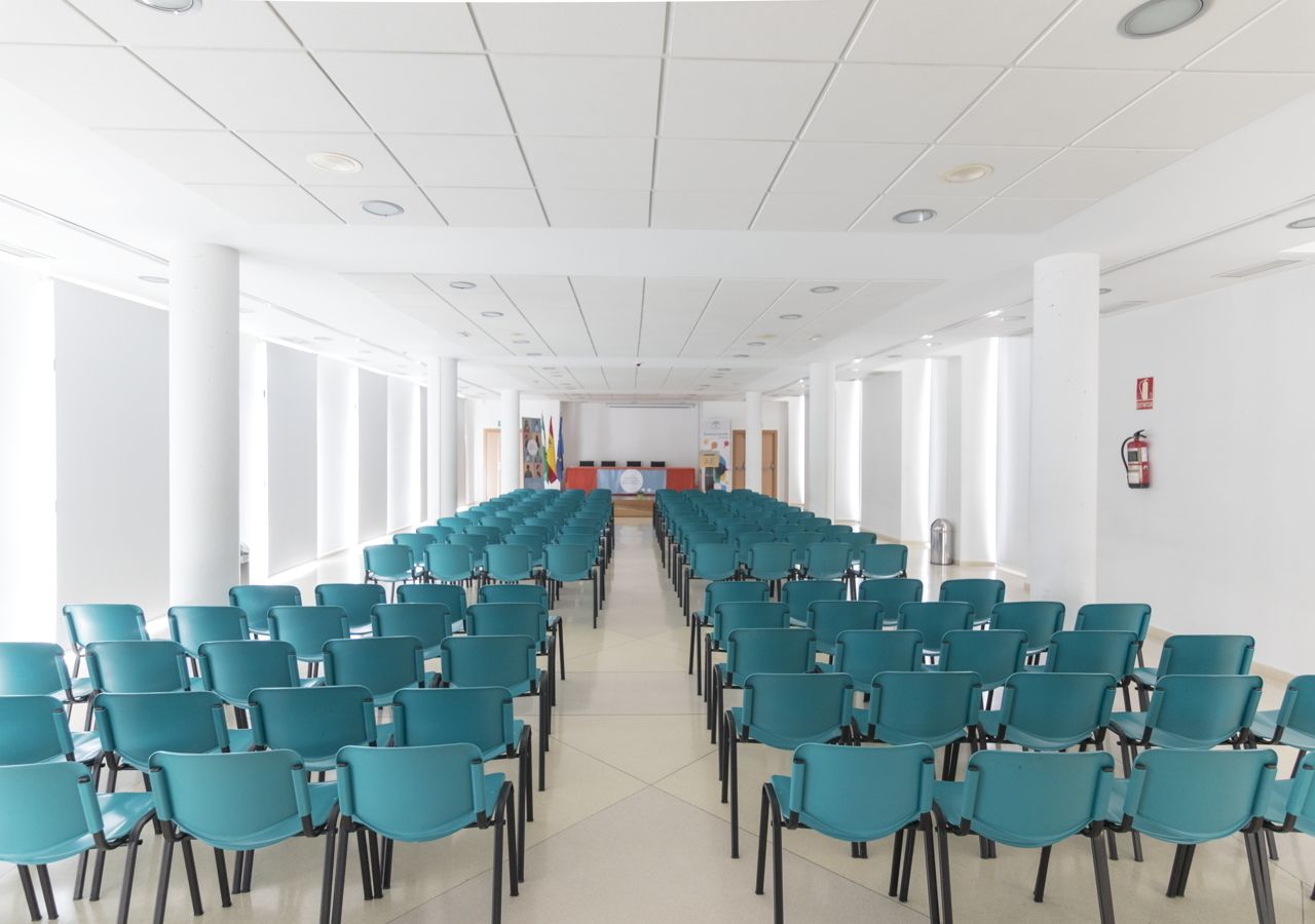 Un salón de conferencias con filas de sillas color turquesa, paredes blancas y un escenario al frente.