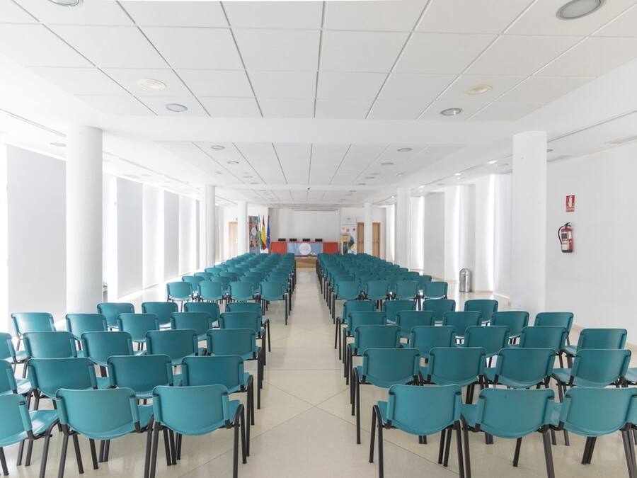 Une salle de conférence avec des rangées de chaises de couleur turquoise, des murs blancs et une scène à l’avant.