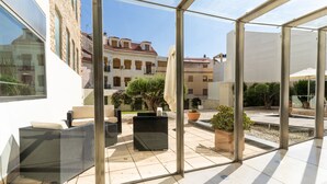 A glass-enclosed patio with outdoor furniture, potted plants, and a view of traditional buildings in the background. - Albergue Inturjoven & Jaen - Hostel (Jaén)