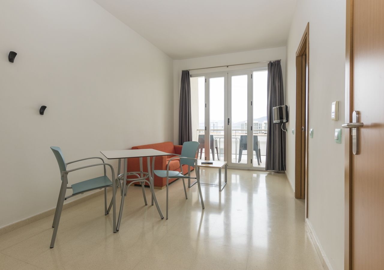 Una habitación de hotel moderna con zona de comedor que incluye una mesa, sillas, un sofá y un amplio ventanal con cortinas.