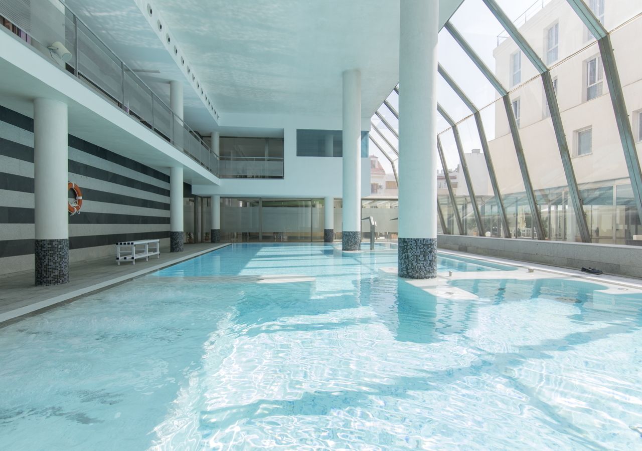 Una piscina interior con agua cristalina azul, rodeada de pilares blancos y un techo de vidrio.