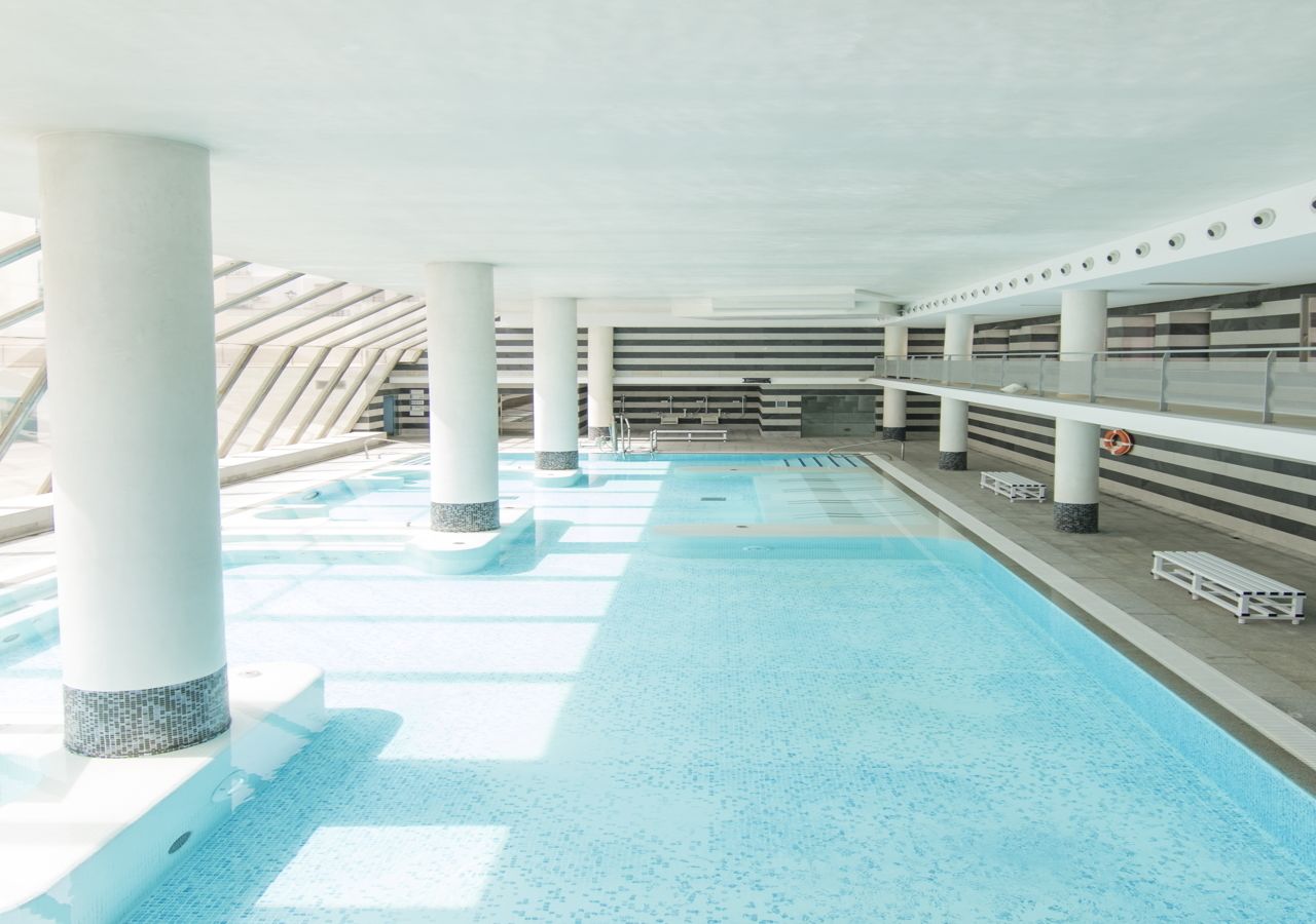 An indoor swimming pool with clear water, white columns, and a tiled floor.
