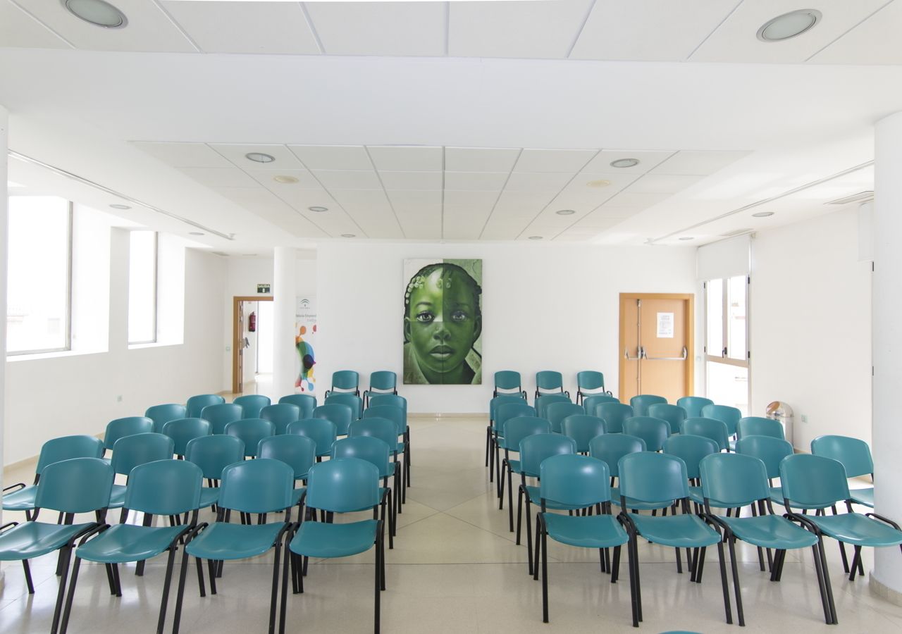 A conference room with rows of blue chairs, a large portrait on the wall, and a door on the right.