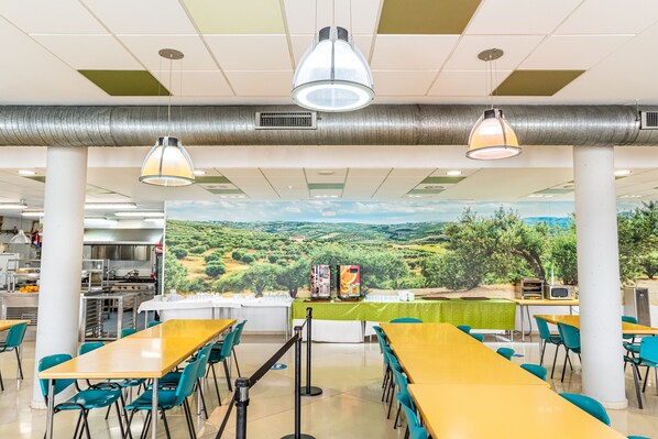 Une cafétéria avec des tables et des chaises, une fresque représentant un paysage et un espace cuisine.