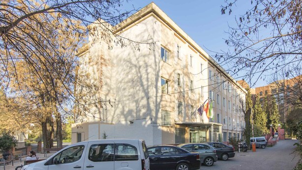 Un edificio de estilo arquitectónico clásico, con una entrada central y varias ventanas. Hay coches aparcados a lo largo de la calle frente al edificio.