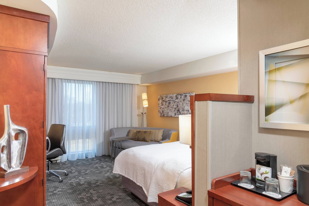 A hotel room with a bed, a desk, a chair, a coffee maker, and a lamp.