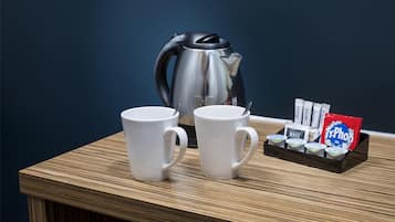 Zwei weiße Tassen, ein schwarzer Wasserkocher und ein Tablett mit Teebeuteln auf einem Holztisch vor dunkler Wand.