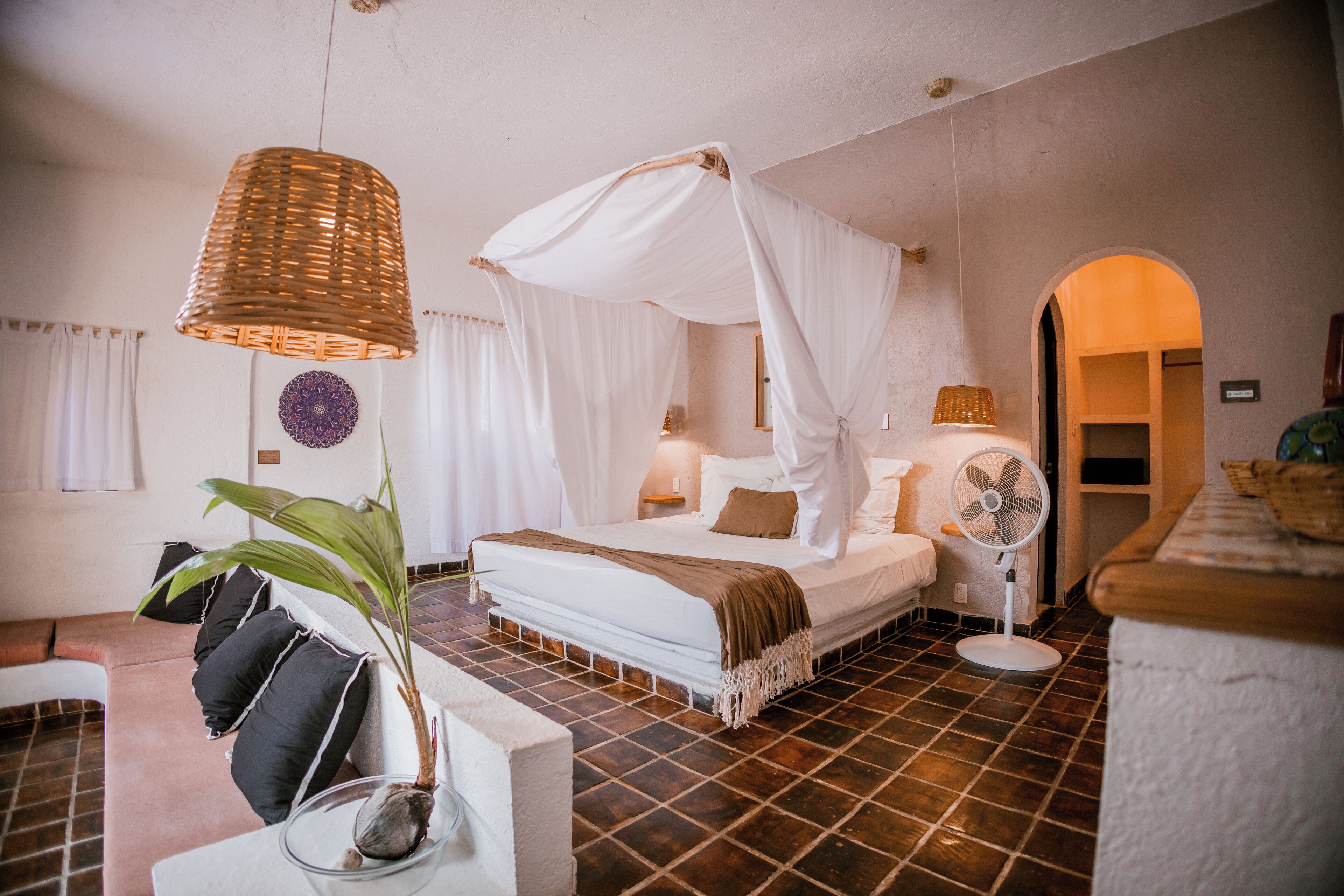 A bedroom with a bed under a canopy, a bedside fan, a potted plant, and a tiled floor.