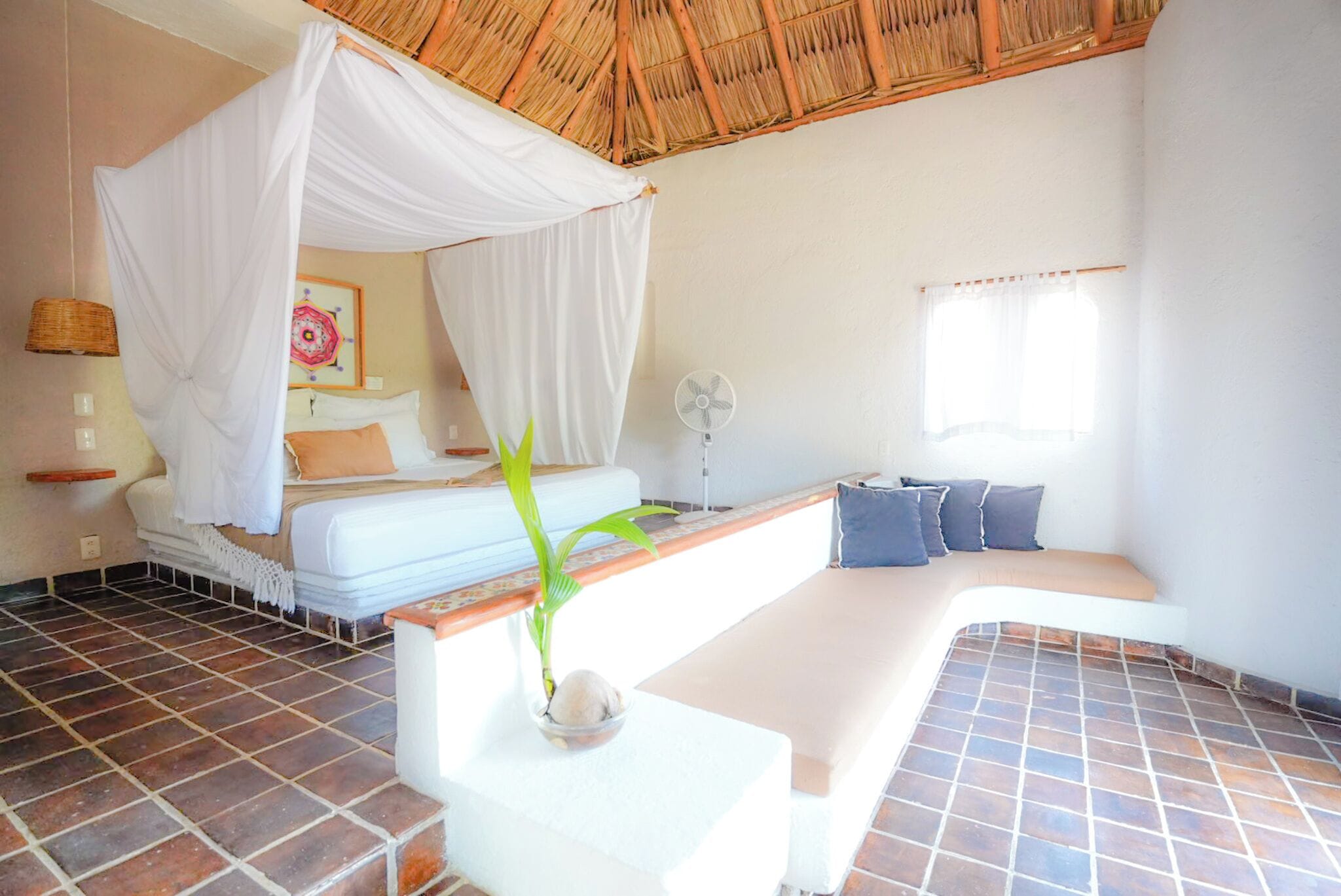 A bedroom with a bed, a daybed, a ceiling fan, a potted plant, and a window with curtains.