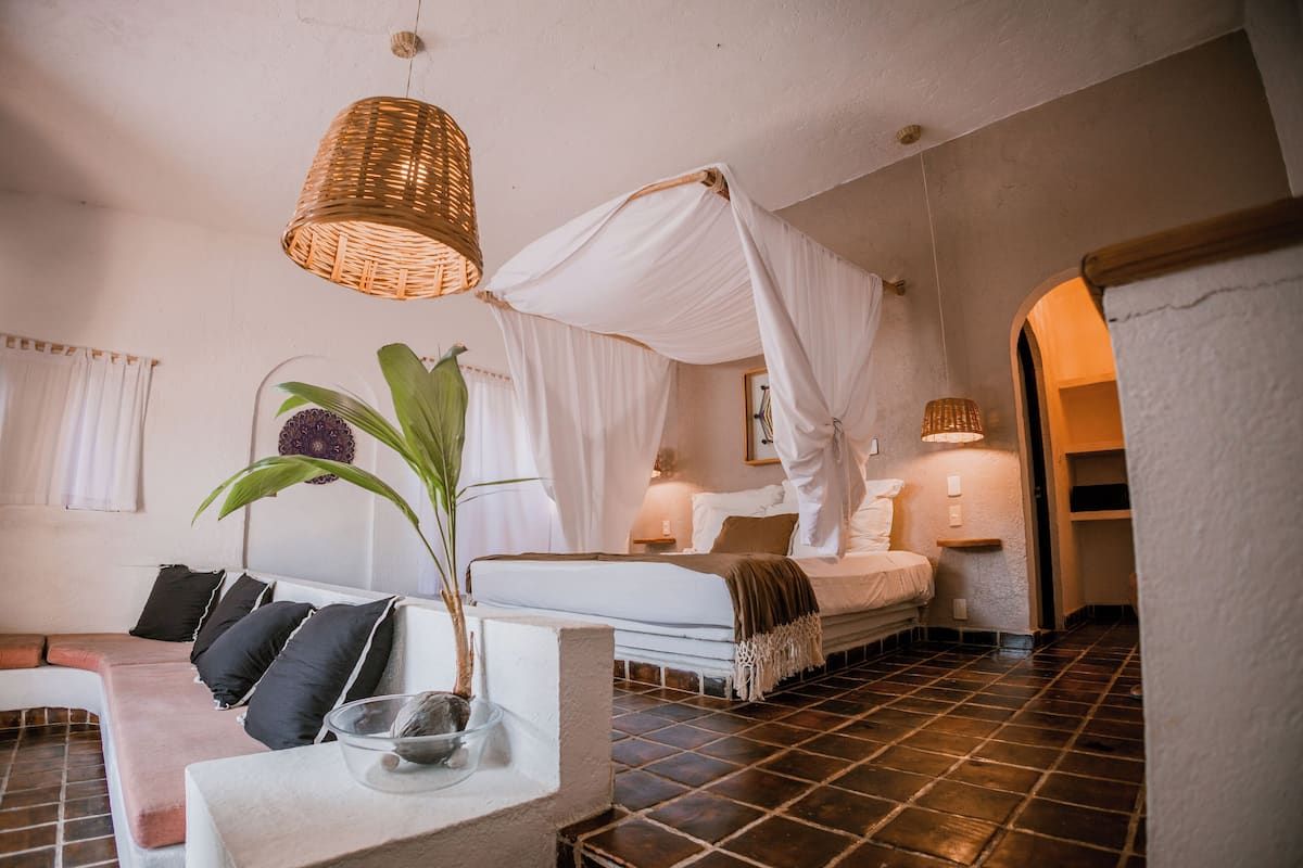 A bedroom with a canopy bed, a bedside table with a plant, and a large window with curtains.