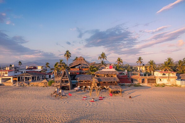 Un complejo turístico frente a la playa con cabañas de techo de paja, palmeras y una vista despejada de las montañas al fondo.