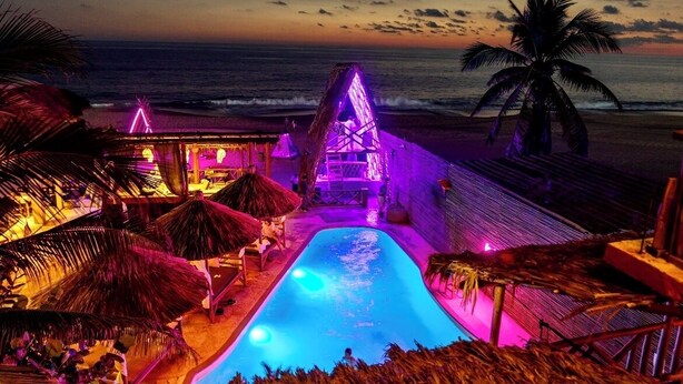 A pool area with a thatched-roof structure, palm trees, and a view of the ocean at sunset. - Hotel BaXar (Acapulco)