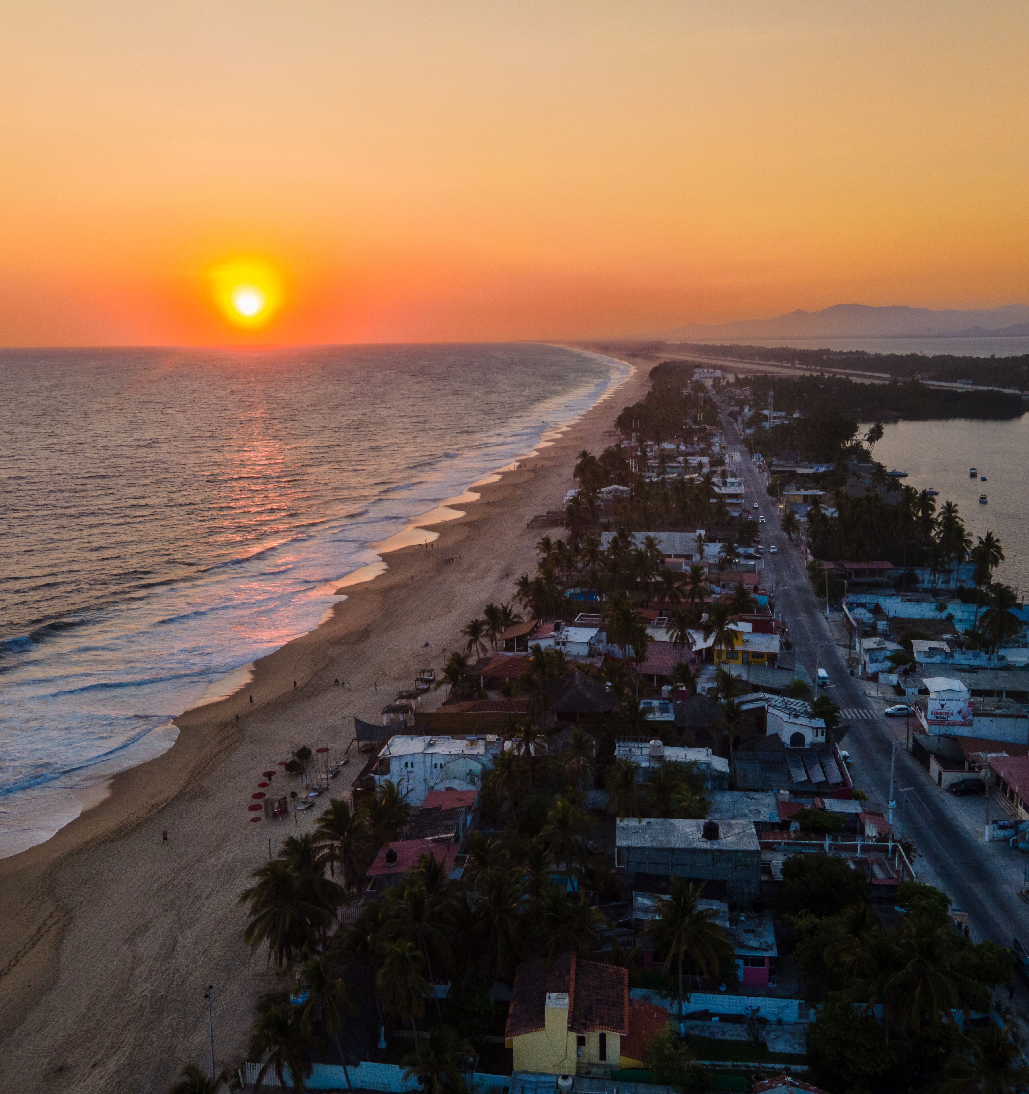 A coastal town at sunset with a beach, houses, and a road leading to the sea.