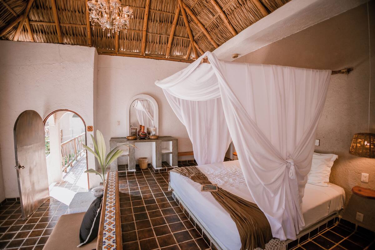 A room with a bed, a desk, a mirror, and a view of the outside.