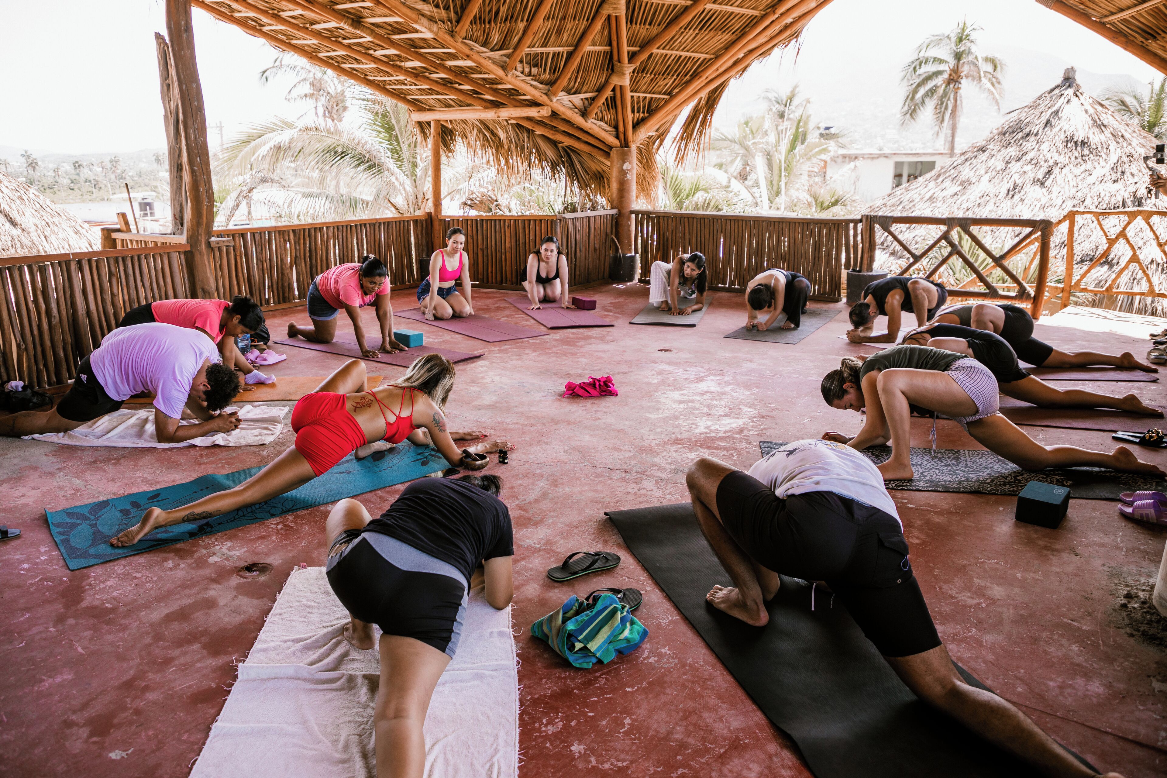 Eine Gruppe von Menschen praktiziert Yoga auf einer überdachten Außenfläche mit einem Reetdach, umgeben von Palmen.