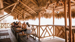 A wooden deck with thatched roof seating and tables overlooking a body of water. - Hotel BaXar (Acapulco)