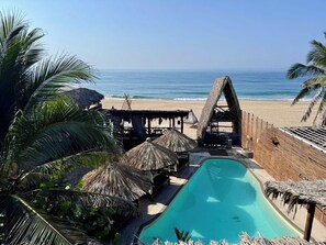 Un área de piscina con vista despejada al océano, rodeada de construcciones de techo de paja y palmeras.
