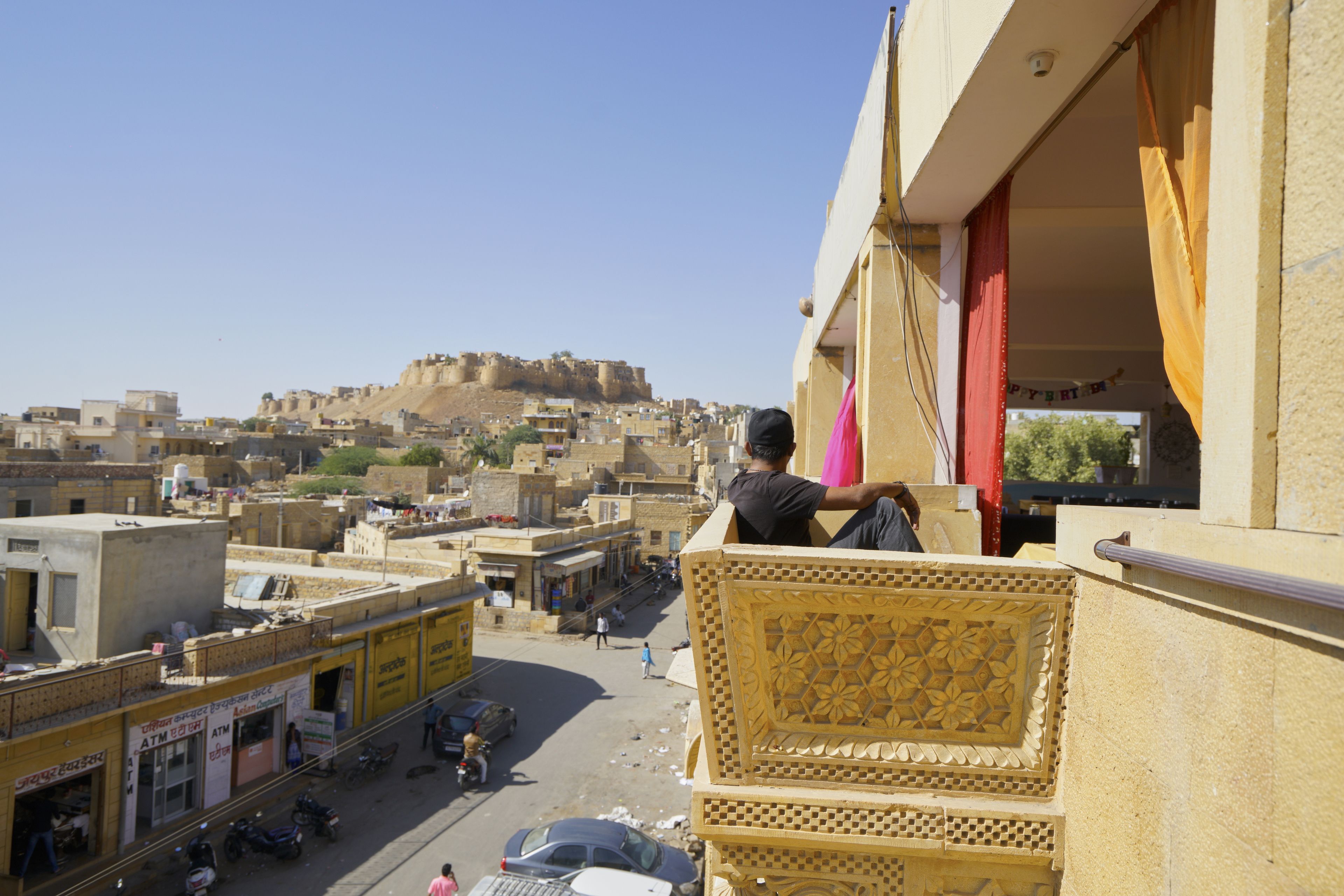 Ein Mann sitzt auf einem Balkon mit Blick auf eine Stadtlandschaft, im Hintergrund eine markante Festung.