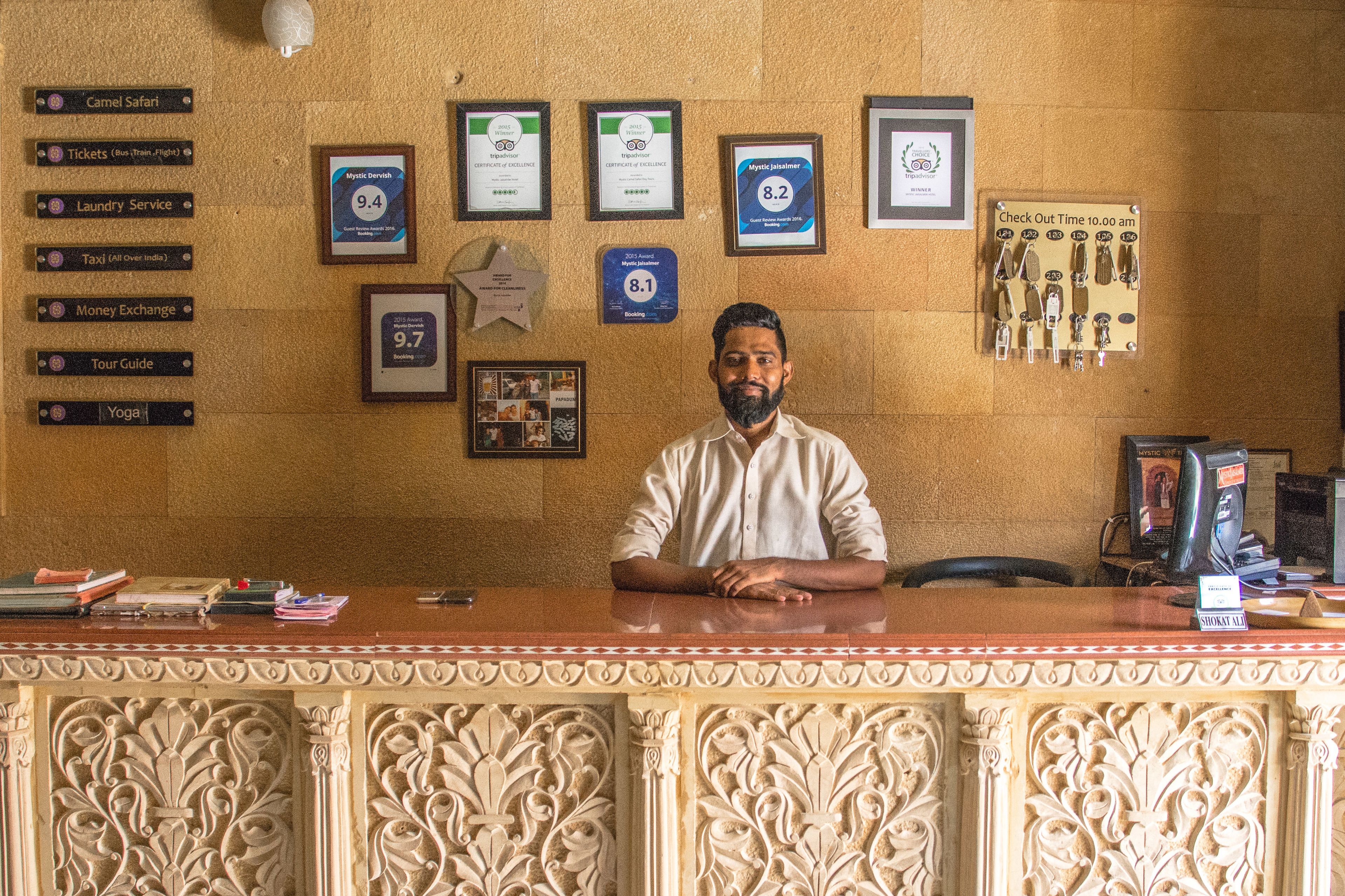 A reception desk with a man standing behind it. The wall behind the desk has various framed certificates and awards, a sign indicating check-out time, and a display of keys.