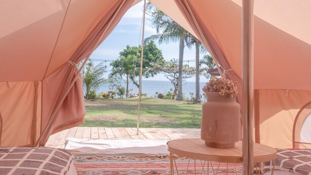 A tent with a view of a grassy area and palm trees, a round table with a vase, and a bed with patterned bedding. - Soka Indah Restaurant & Bungalows (Selemadeg)