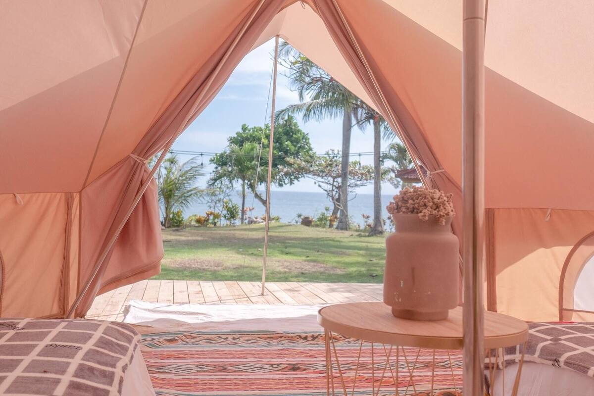 A tent with a view of a grassy area and palm trees, a round table with a vase, and a bed with patterned bedding.