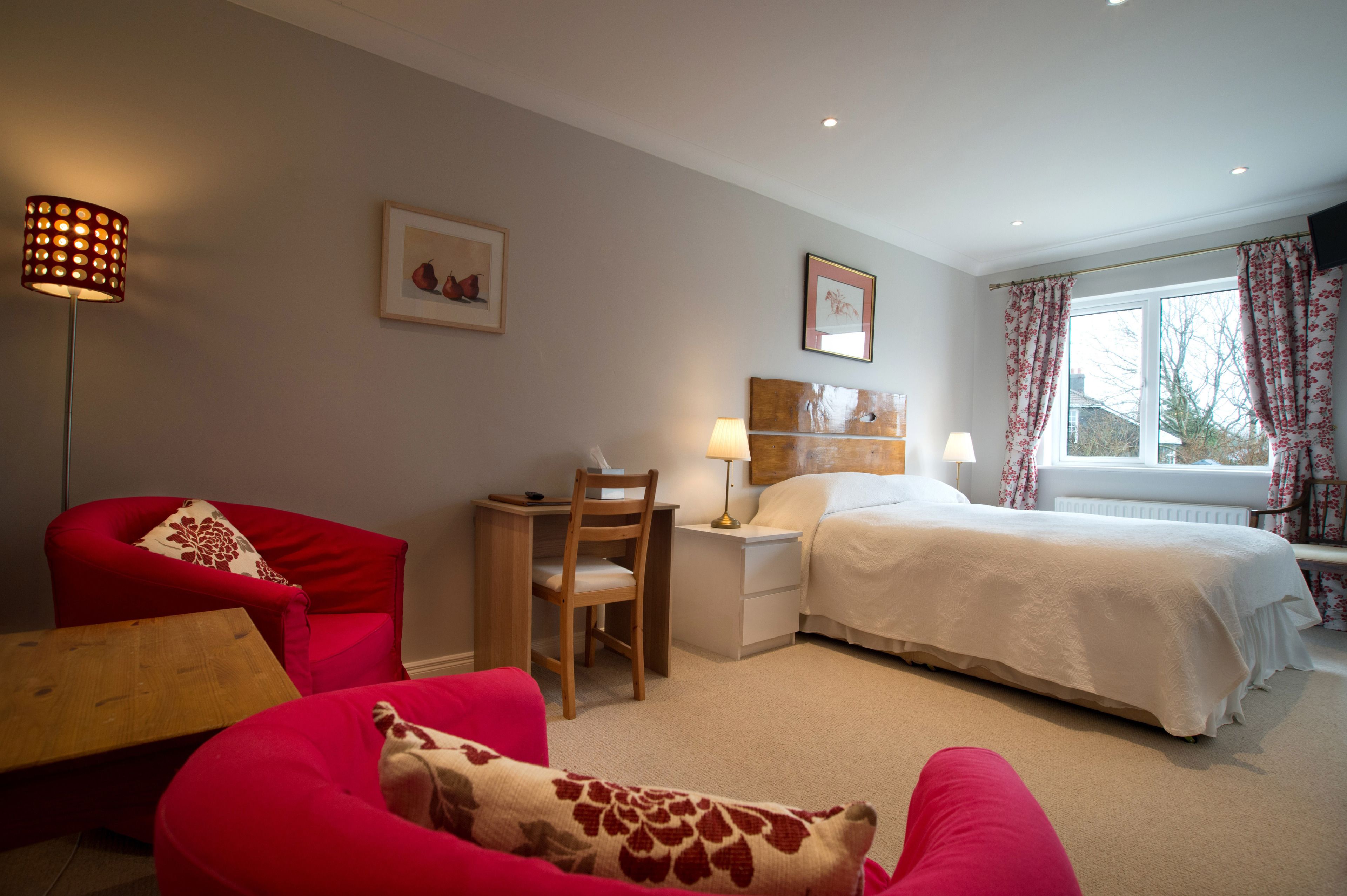 A bedroom with a bed, a desk, a chair, a red armchair, and a lamp.