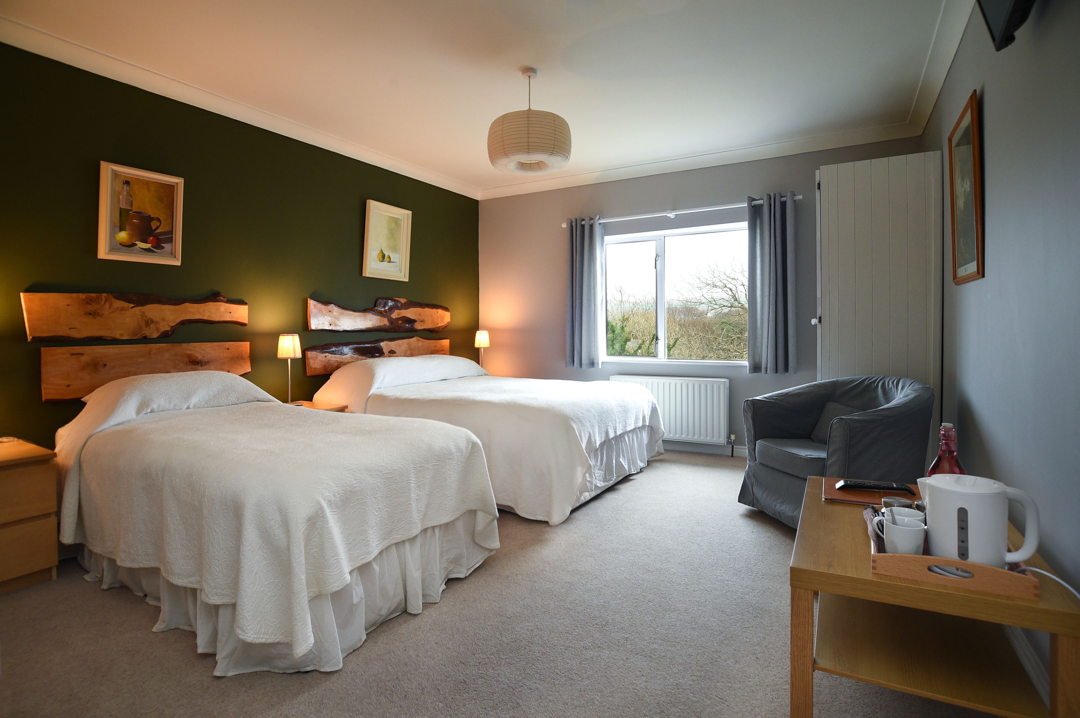 A bedroom with two beds, a wooden headboard, a window with curtains, a radiator, a grey sofa, and a bedside table with a kettle and mug.