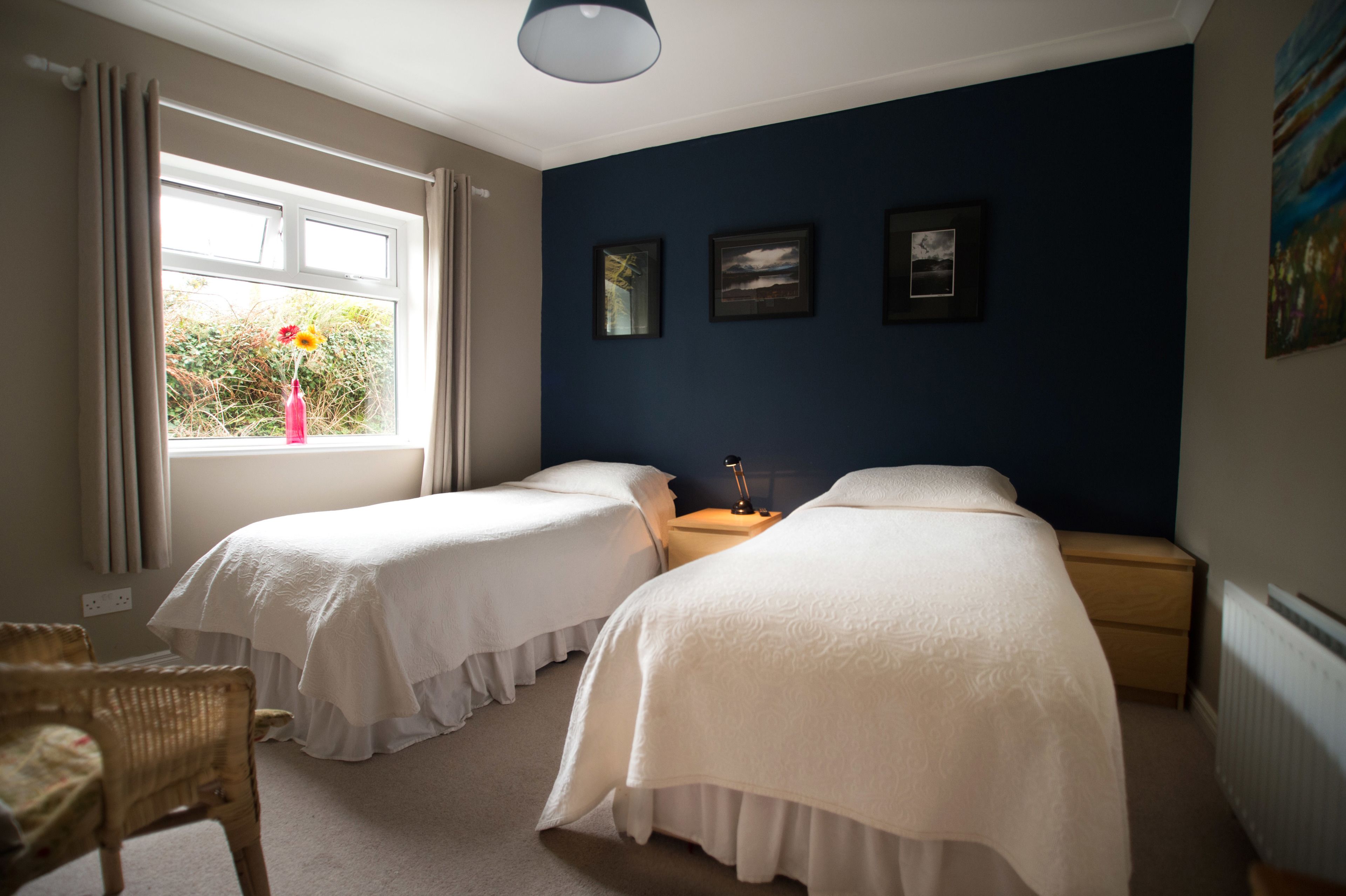 A bedroom with two beds, a window with curtains, a chair, and framed pictures on the wall.