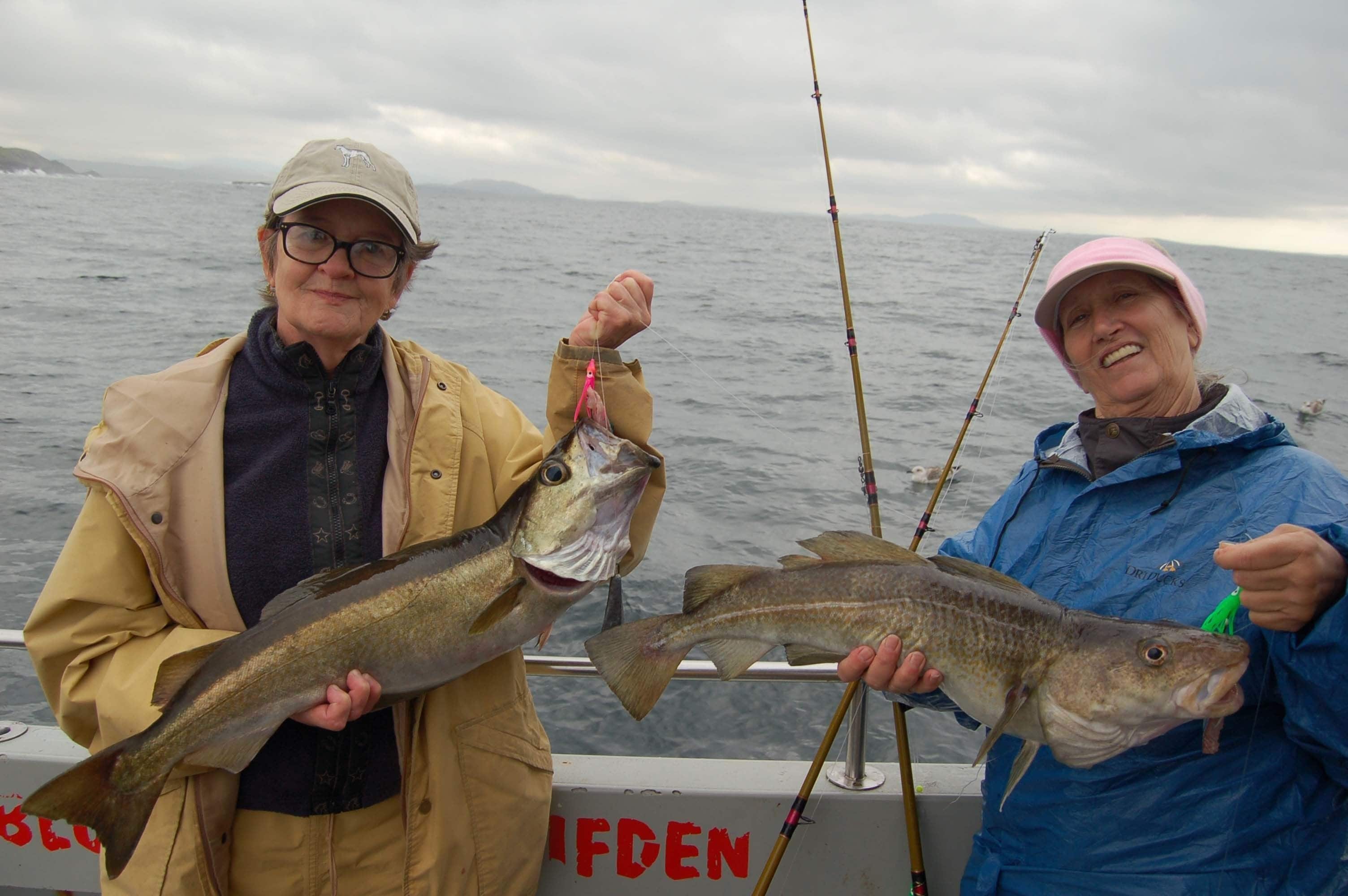 Zwei Personen halten Fische auf einem Boot, im Hintergrund ein Gewässer und Berge.