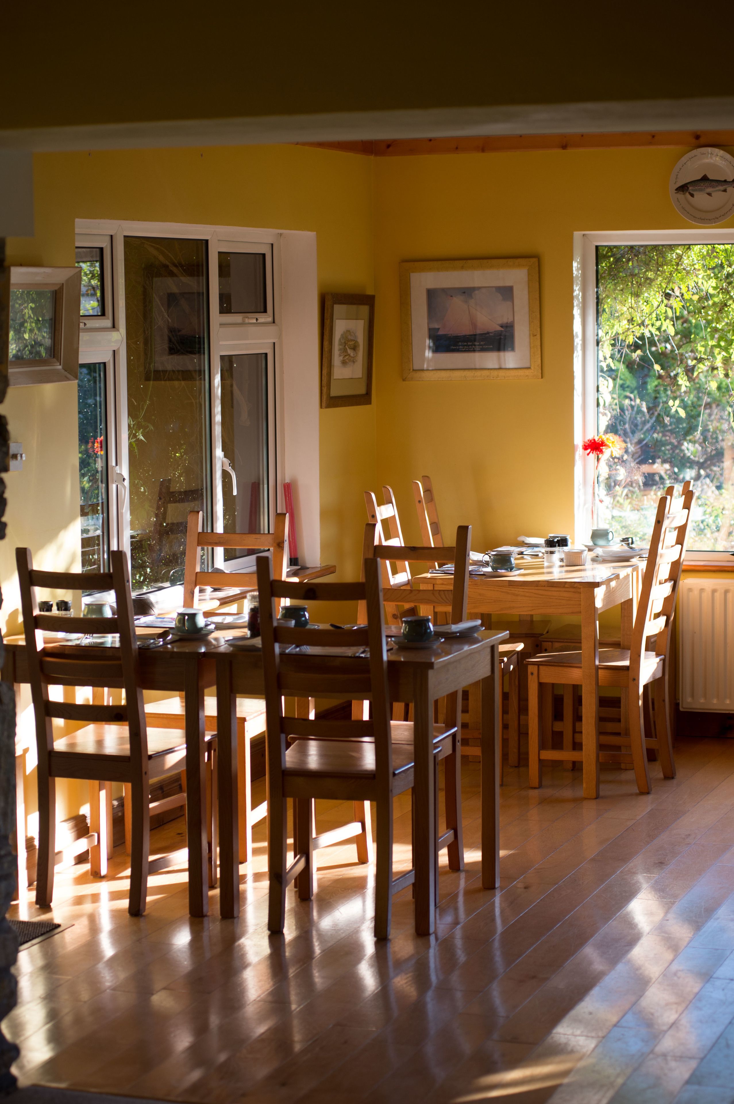 A dining area with wooden furniture, a table, and chairs. There are windows with blinds, a radiator, and a wall-mounted clock.