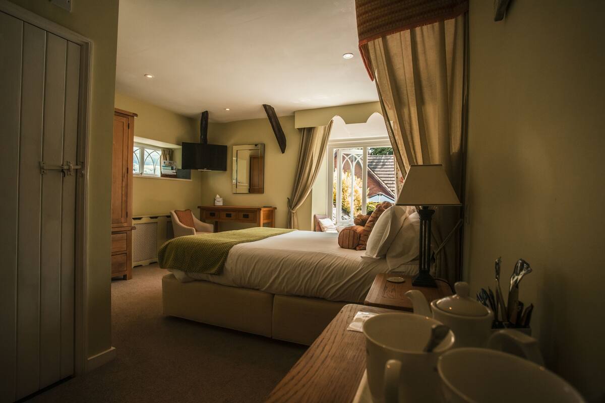 Quarto com cama, mesa de cabeceira, luminária e janela com cortinas.