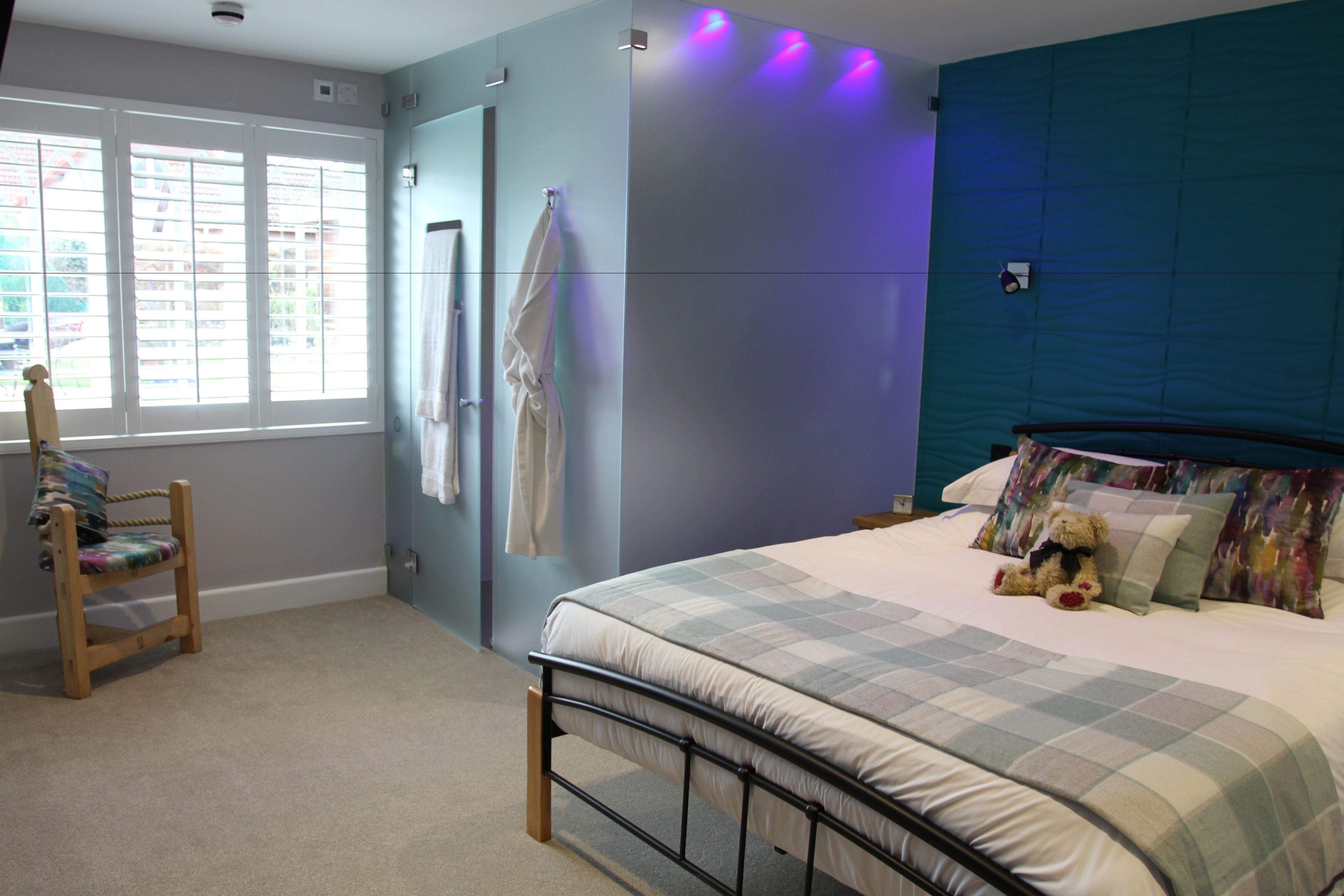 Un dormitorio con cama, una silla, un ventanal y una pared con lámpara.