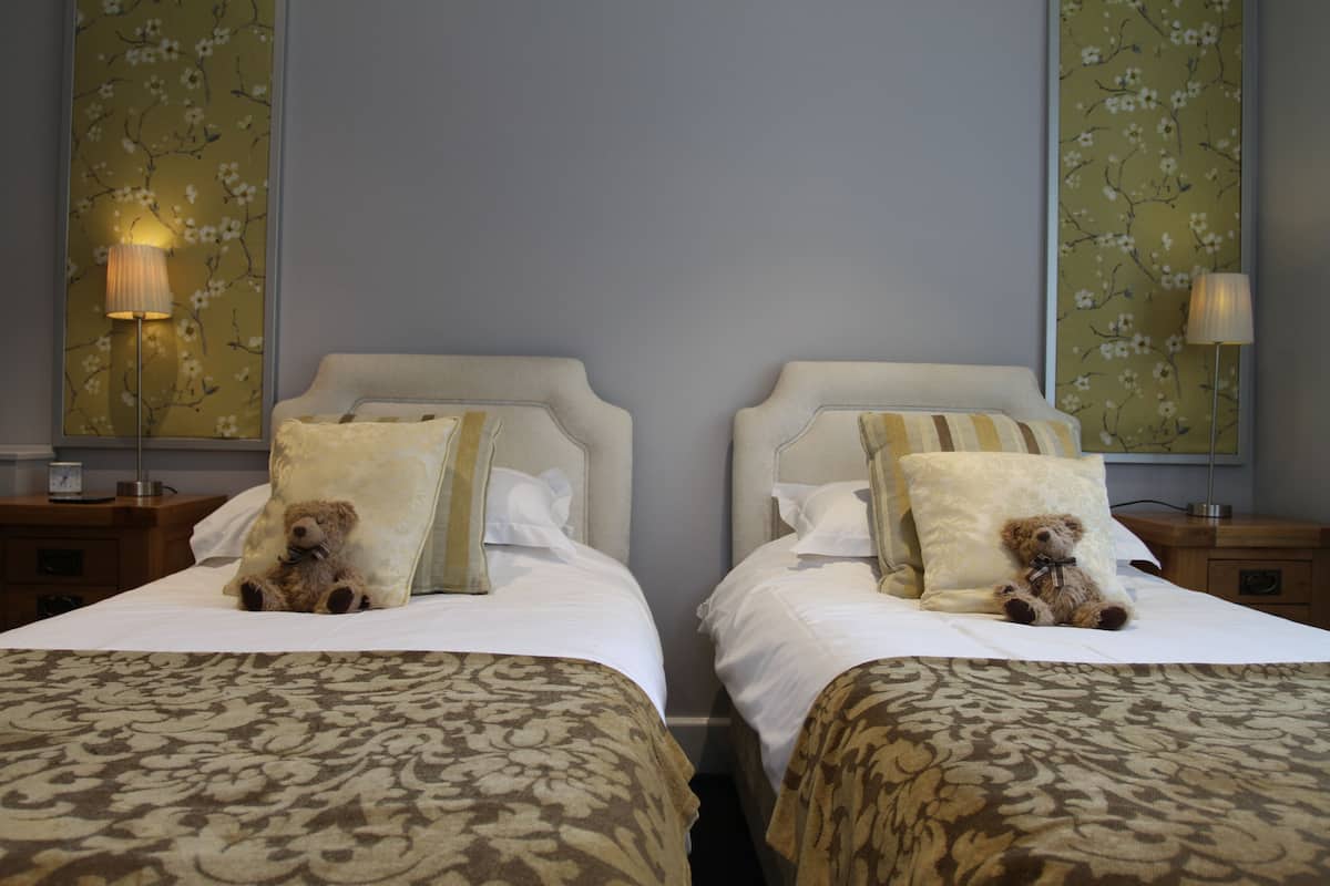 Quarto de hotel com duas camas, cada uma com um urso de pelúcia, uma luminária e papel de parede floral.