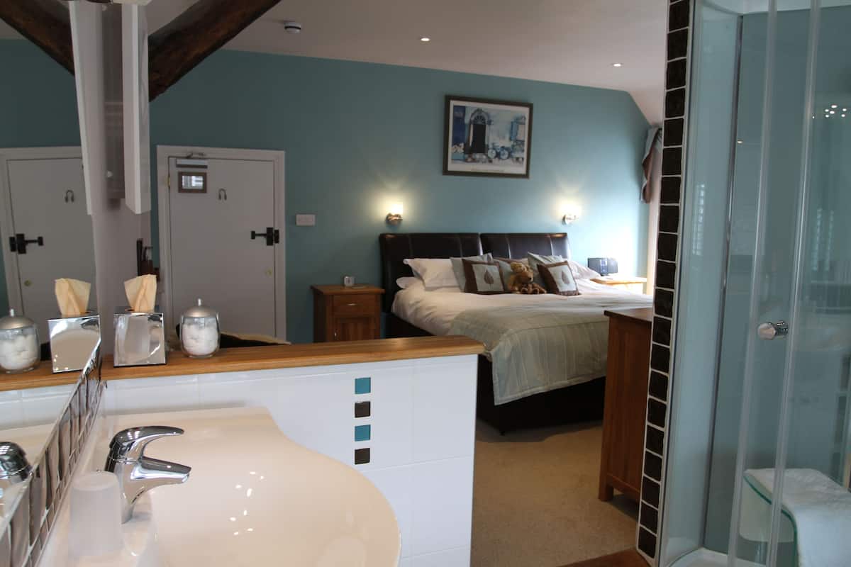 Quarto de hotel com uma grande pia no banheiro, uma cama com cabeceira de madeira, box de vidro para o chuveiro e um quadro na parede.