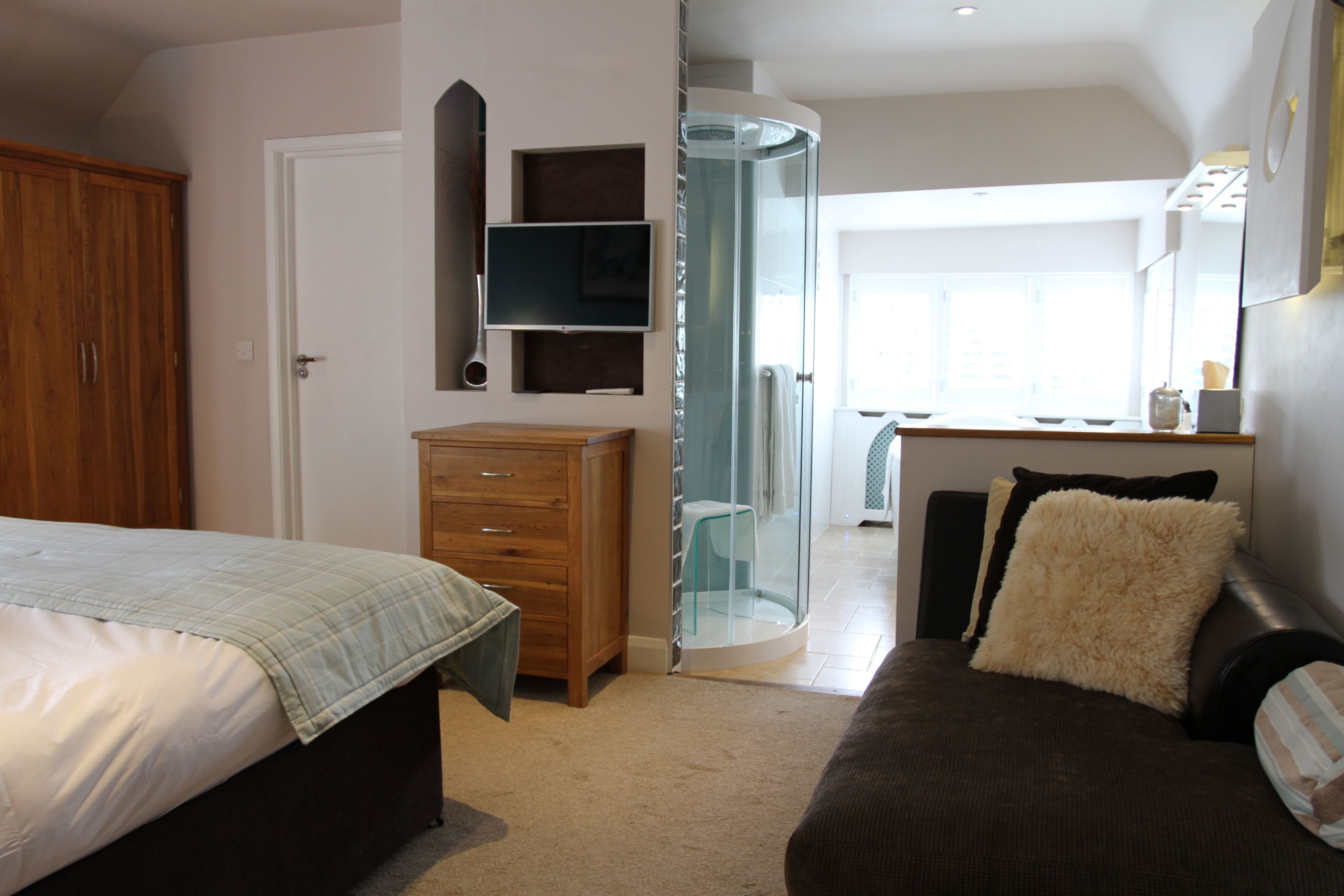 A bedroom with a bed, a wooden dresser, a glass shower enclosure, and a desk with a chair.