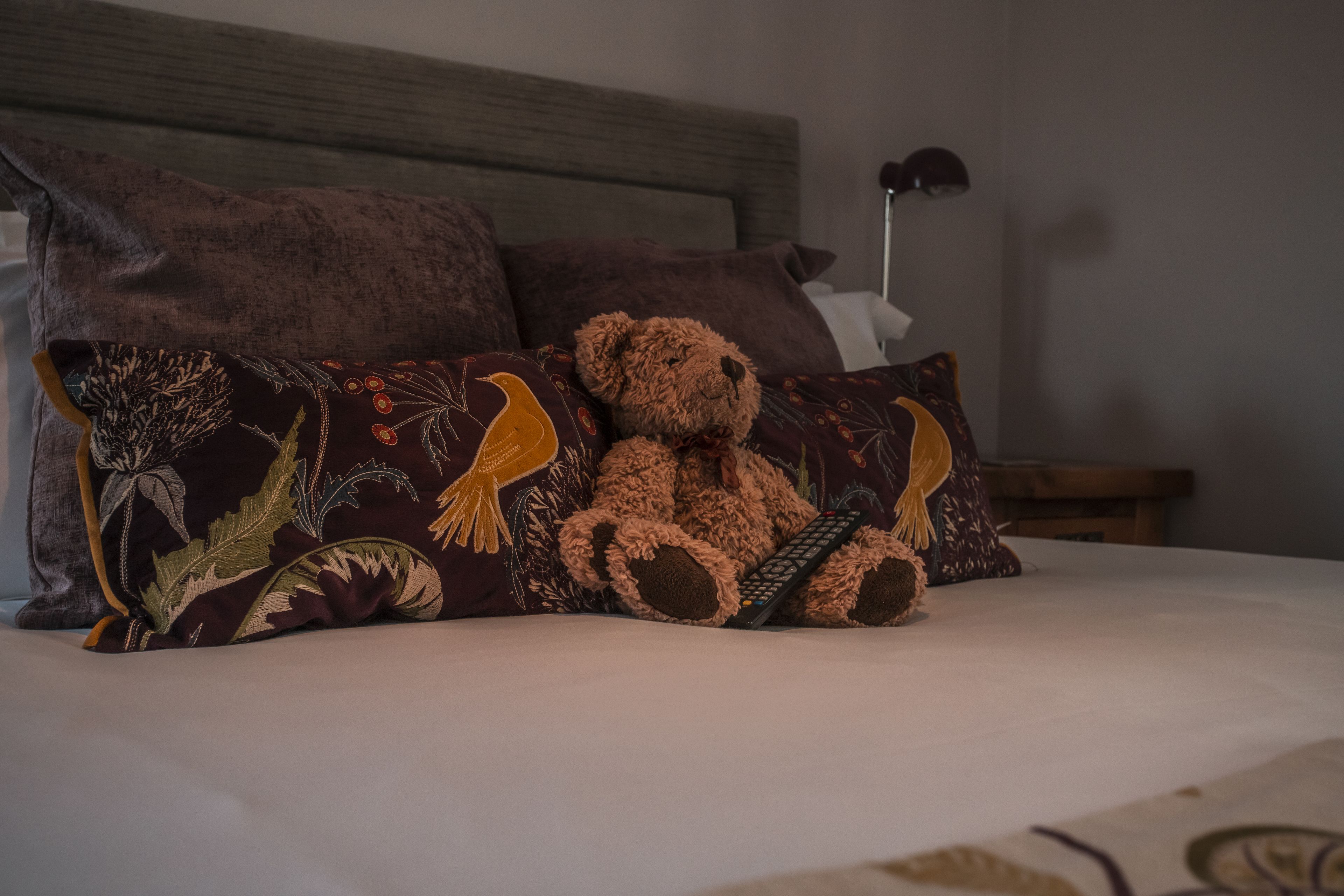 A bed with a teddy bear and a remote control, surrounded by pillows with floral patterns.