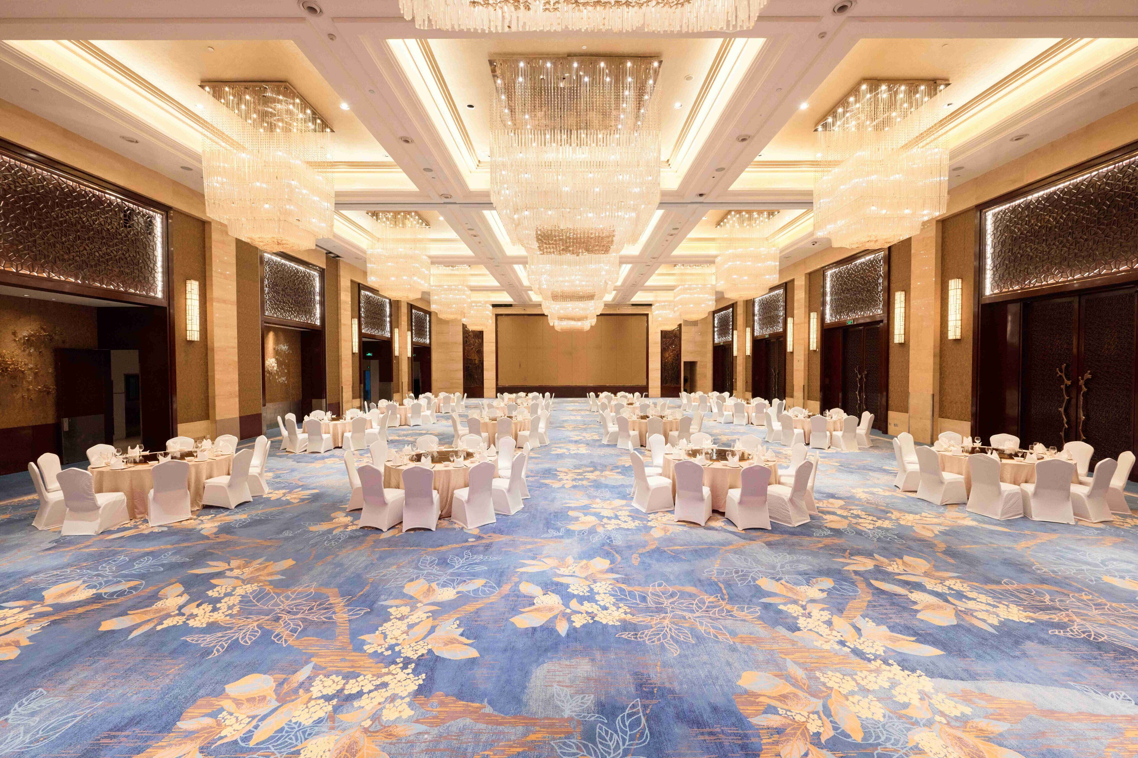 Un amplio salón de banquetes con filas de mesas y sillas, una alfombra azul con motivos florales y candelabros ornamentados.